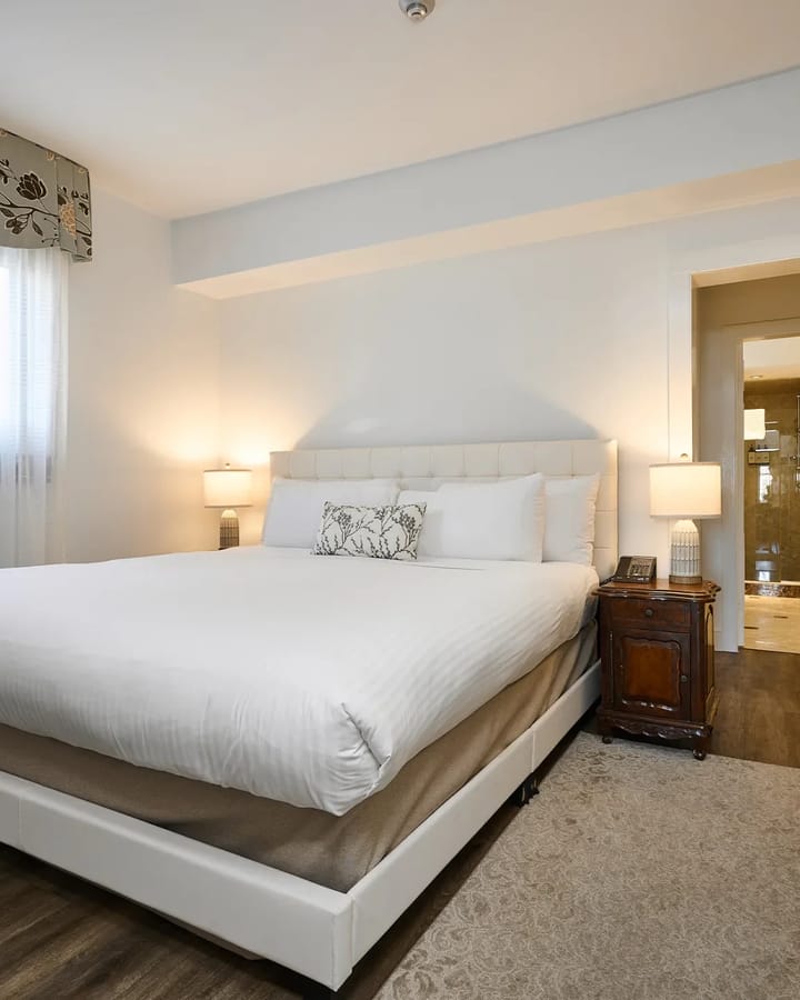 A bright hotel guest room with a made king bed, bedside lamps and an open door to the ensuite