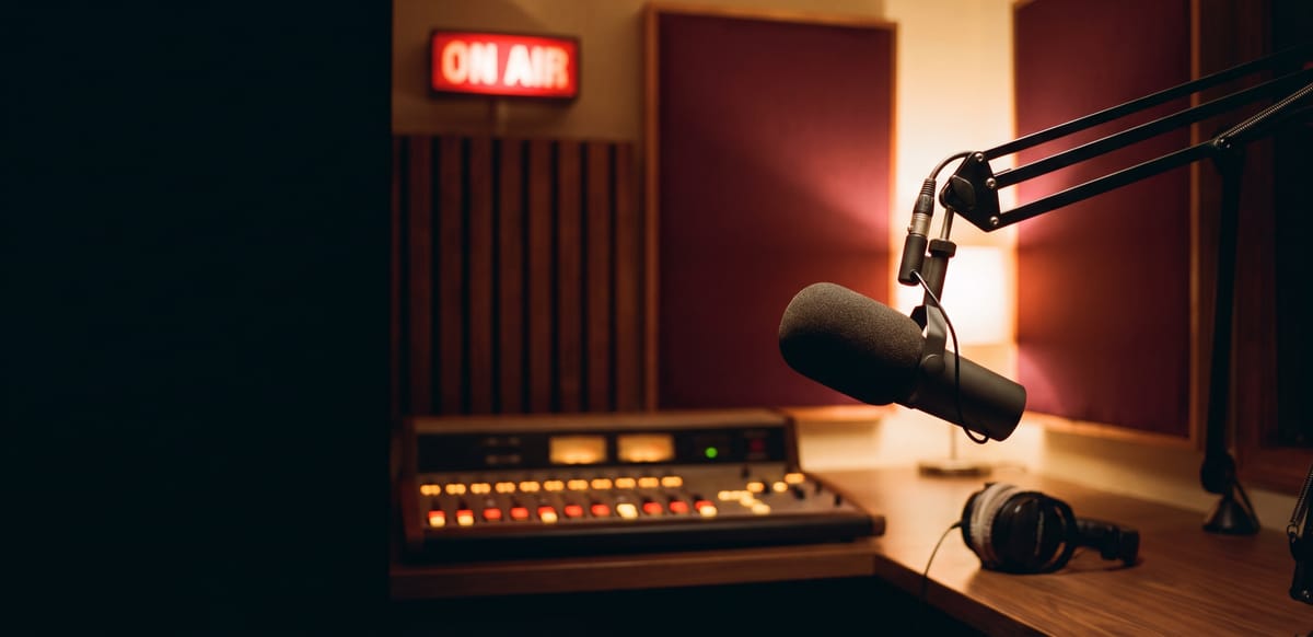 A broadcast radio microphone in a warm, dimly lit studio with an On Air light glowing behind it