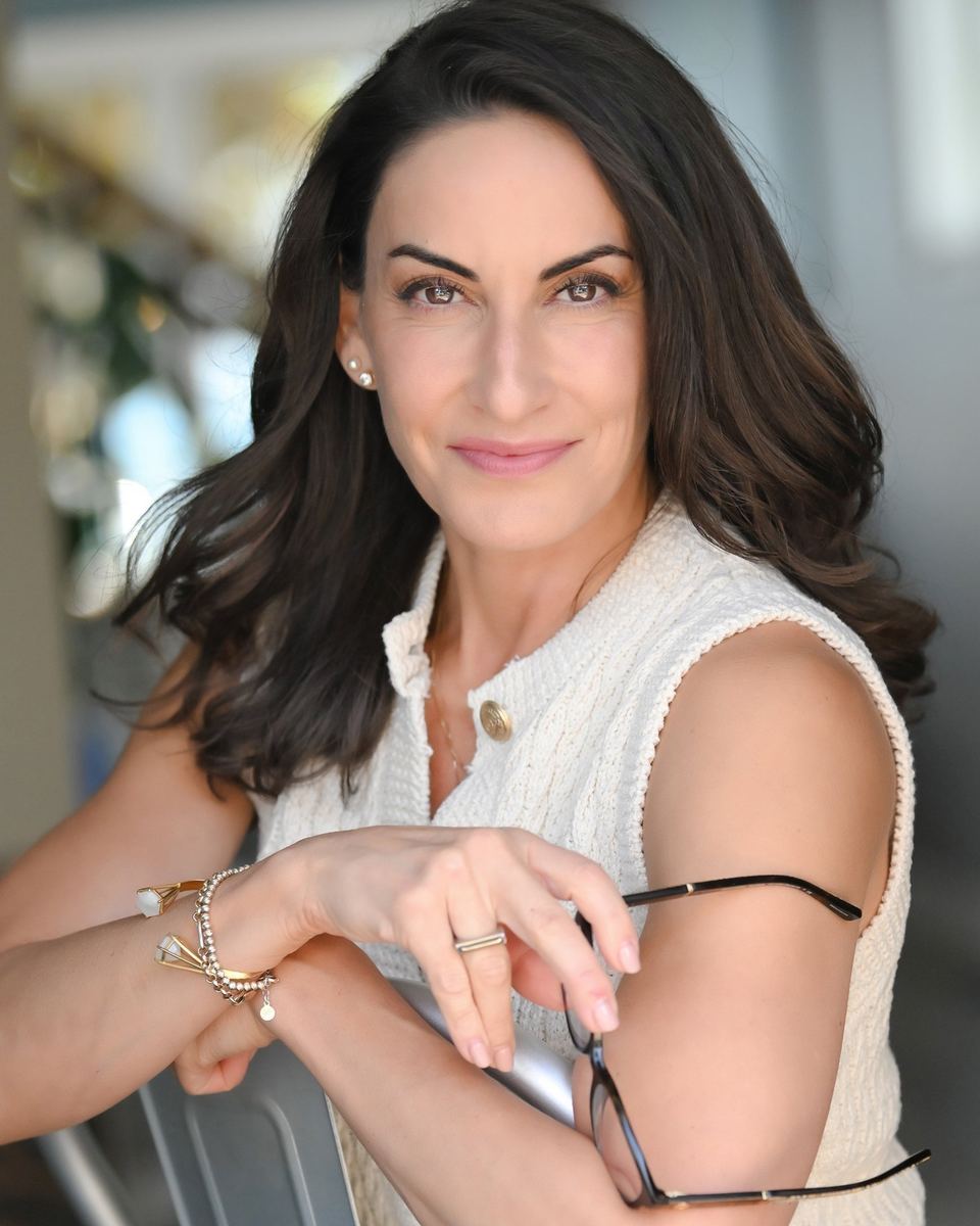Kim Rossi in a cream knit top, leaning on a chair and holding her glasses