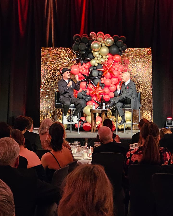 Howie Mandel in conversation on stage at the Hope Gala, in front of a coral, black and gold balloon wall